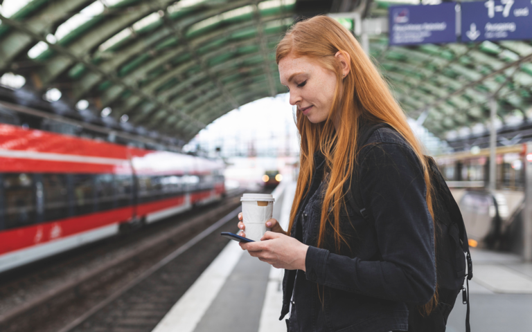 Junge Frau auf dem Weg zur Arbeit mit Kaffee to go und Smartphone auf dem Bahnsteig eines Berliner Bahnhofs