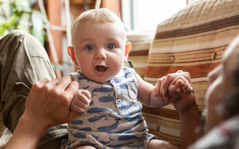 Playful baby sitting with father at home, Portrait of cheerful and excited infant baby sitting on father lying on couch and holding hands while playing and jumping on stomach, Symbolfoto