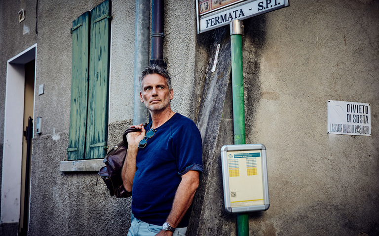 Oliver Stock wartet mit Reisetasche in einem italienischen Dorf an einer Bushaltestelle