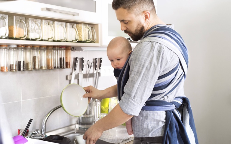 Vater mit Baby im Tragetuch zu Hause beim Abwaschen (Symbolbild)