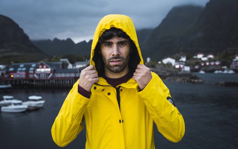 Junger Mann mi Bart in gelber Regenkapuzenjacke vor Fischersiedlung auf den Lofoten, Norwegen